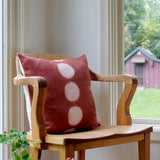 Hand dyed itajime kamppinen moon rust throw pillow with 5 undyed circles down middle on wooden chair with windows on both sides