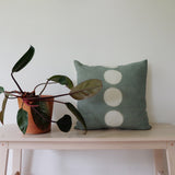 Sage handdyed hemp linen 18" square pillow with 5 undyed circles in vertical center line. Sits on bench with plant. Made by Kamppinen